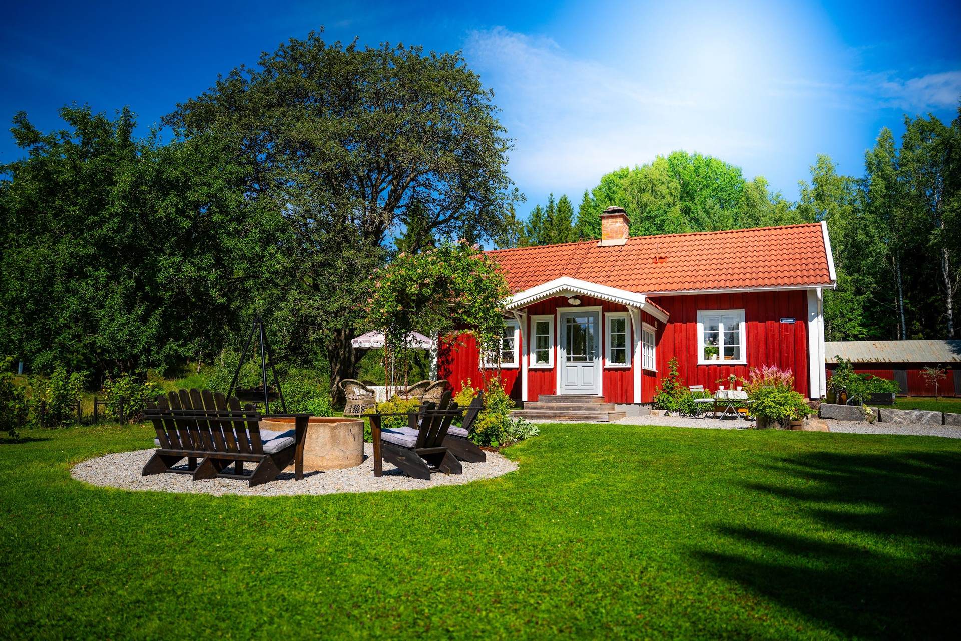 The red timber house of Djäknetuva with its lawn, fire pit and forest behind, on a summer day