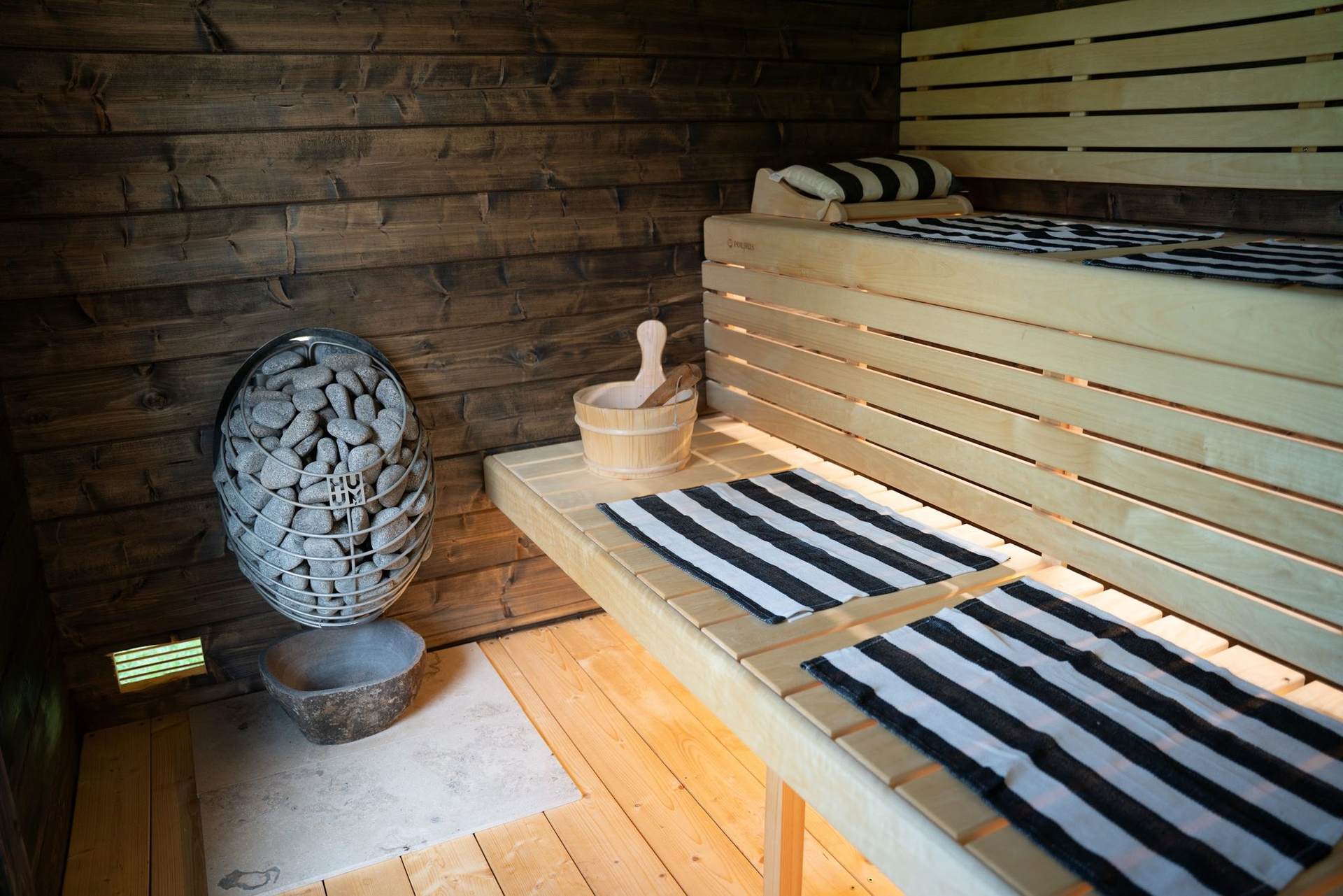 Inside the sauna: pale wooden benches with striped towels, a stone-filled heater and a wooden bucket